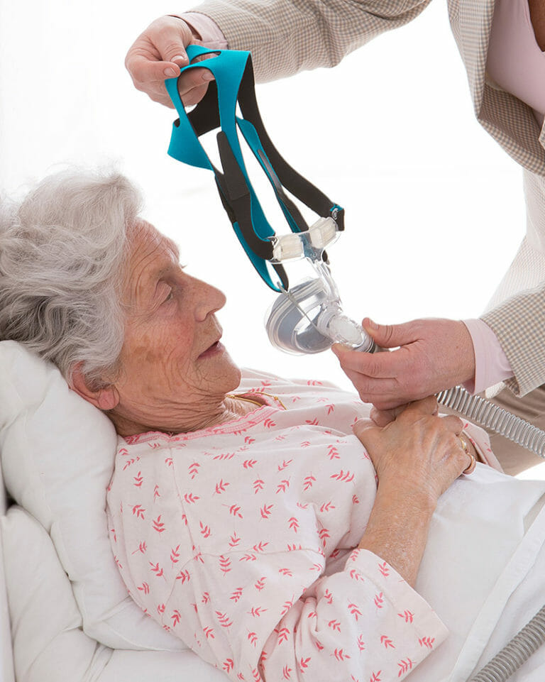 A caregiver helps an elderly woman lying in bed by placing a CPAP mask over her face