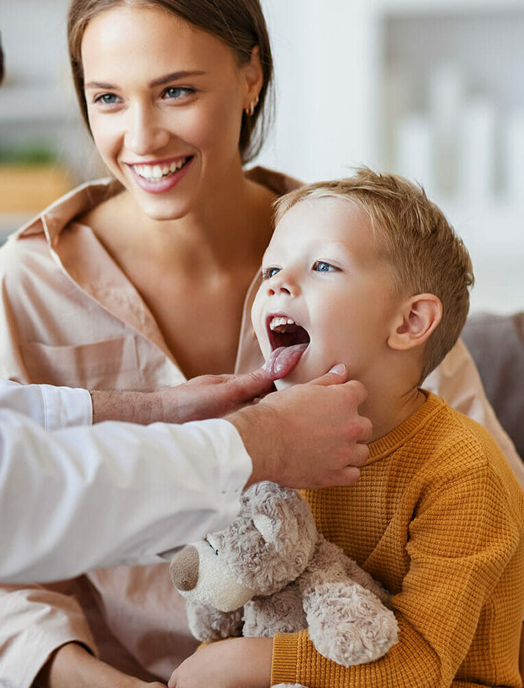 A smiling mother holds her son as a doctor checks the child’s open mouth during a medical exam