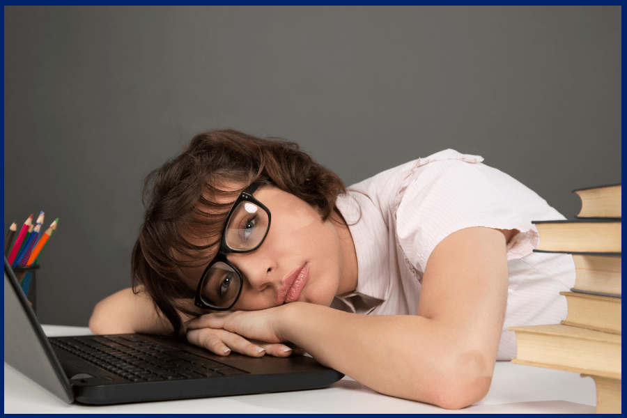 Tired student experiencing study burnout, resting head on laptop with books and pencils nearby in a classroom setting.
