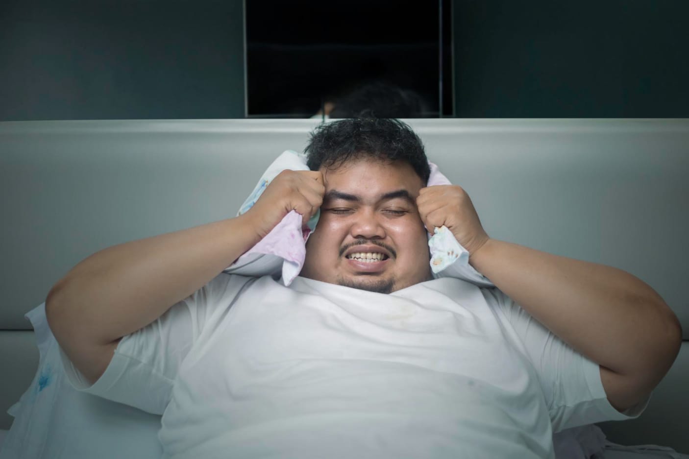 Man in bed covering his ears with a pillow, appearing frustrated