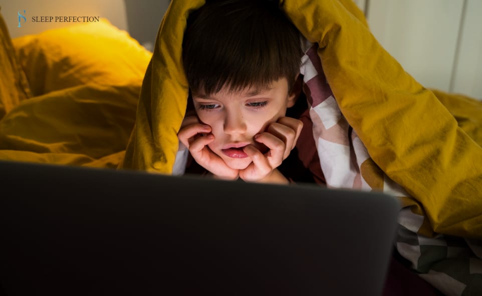 Child lying under a blanket while watching a screen at night