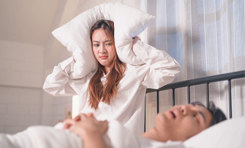 A frustrated woman covers her ears with a pillow while her partner snores loudly beside her in bed
